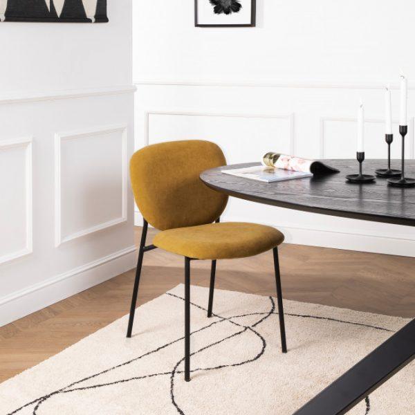 La chaise présente un rembourrage de couleur jaune moutarde, qui peut évoquer la sensation d'une étoffe douce avec une teinte chaude et profonde, semblable à la couleur d'une épice douce ou d'une moutarde ancienne. L'assise et le dossier semblent généreusement garnis, suggérant un confort moelleux. Les lignes du dossier légèrement courbé et de l'assise sont simples et épurées. La structure de la chaise est métallique avec des pieds fins et droits, peints en noir, offrant un contraste élégant avec le jaune de l'assise. Ces pieds dénotent une silhouette à la fois moderne et discrète, qui pourrait facilement s'intégrer dans divers styles de décoration intérieure. L'ensemble crée une pièce de mobilier à l’esthétique contemporaine et chaleureuse.